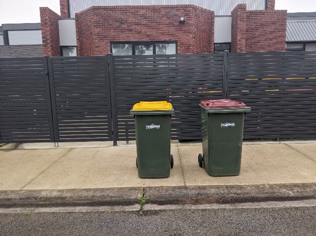 Two bins side by side on a footpath in front of a fence.
